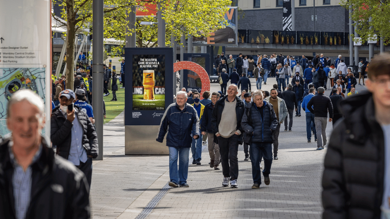 Crowd of people walking along a wide pedestrian area lined with trees and street poles, with some individuals wearing jackets and some wearing face masks, and a digital advertisement promoting Carling beer visible among the poles.