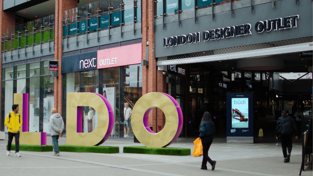 Outdoor shopping area with large decorative letters spelling 'LDO' on grass patches, people walking nearby, storefronts including Next Outlet and London Designer Outlet visible in the background.