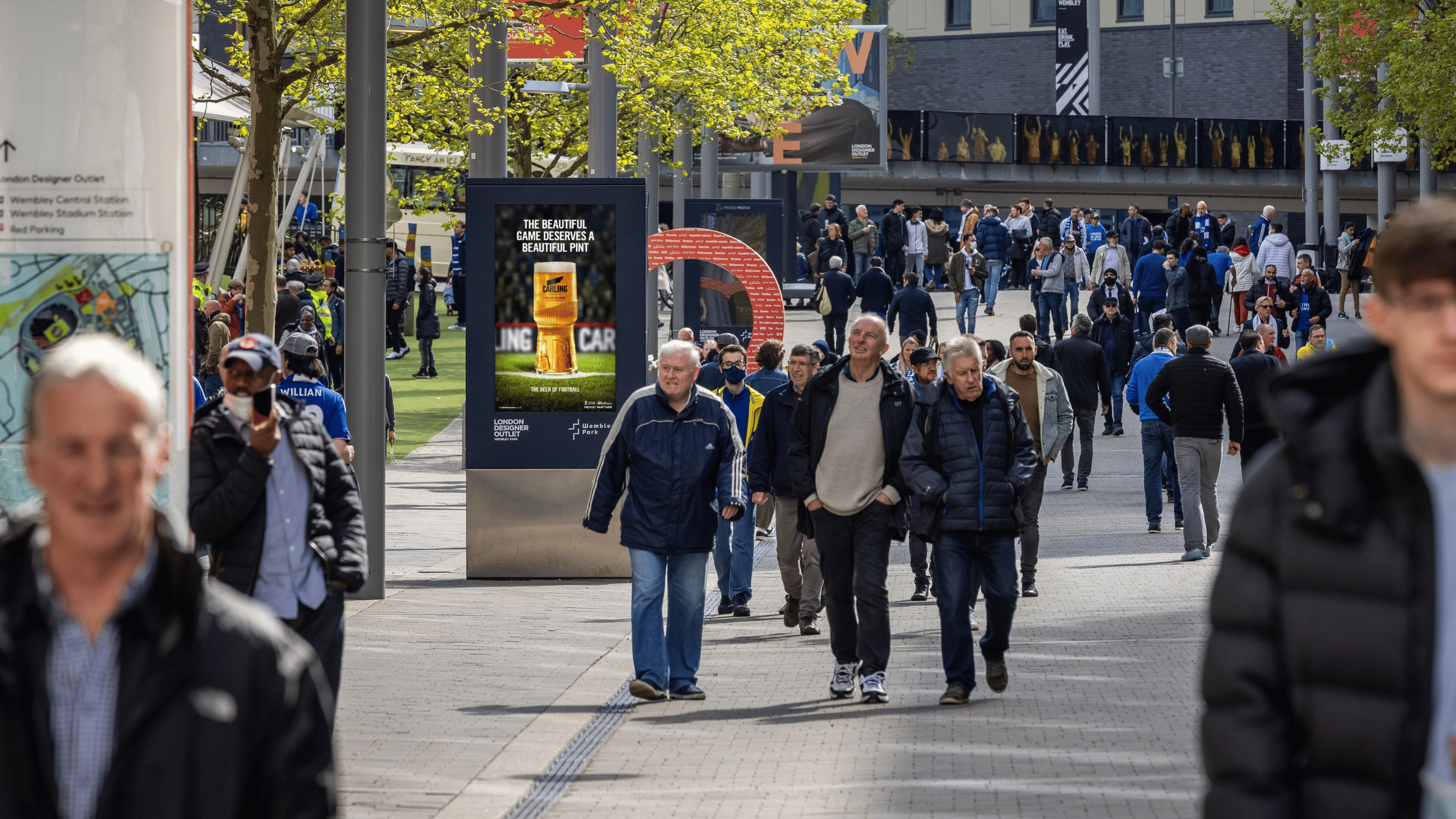 Crowd of people walking along a wide outdoor pathway lined with trees and advertising boards, with most wearing jackets on a sunny day.