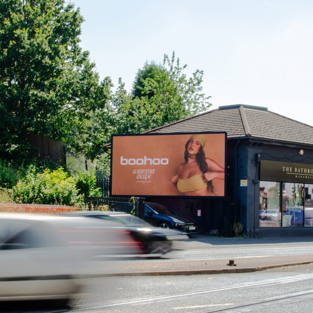 Outdoor billboard on a building corner advertising Boohoo with a photo of a woman wearing a yellow bandeau top and head wrap, text reading 'a summer escape'. Motion-blurred cars pass on the street in front. Lush green trees frame the scene.