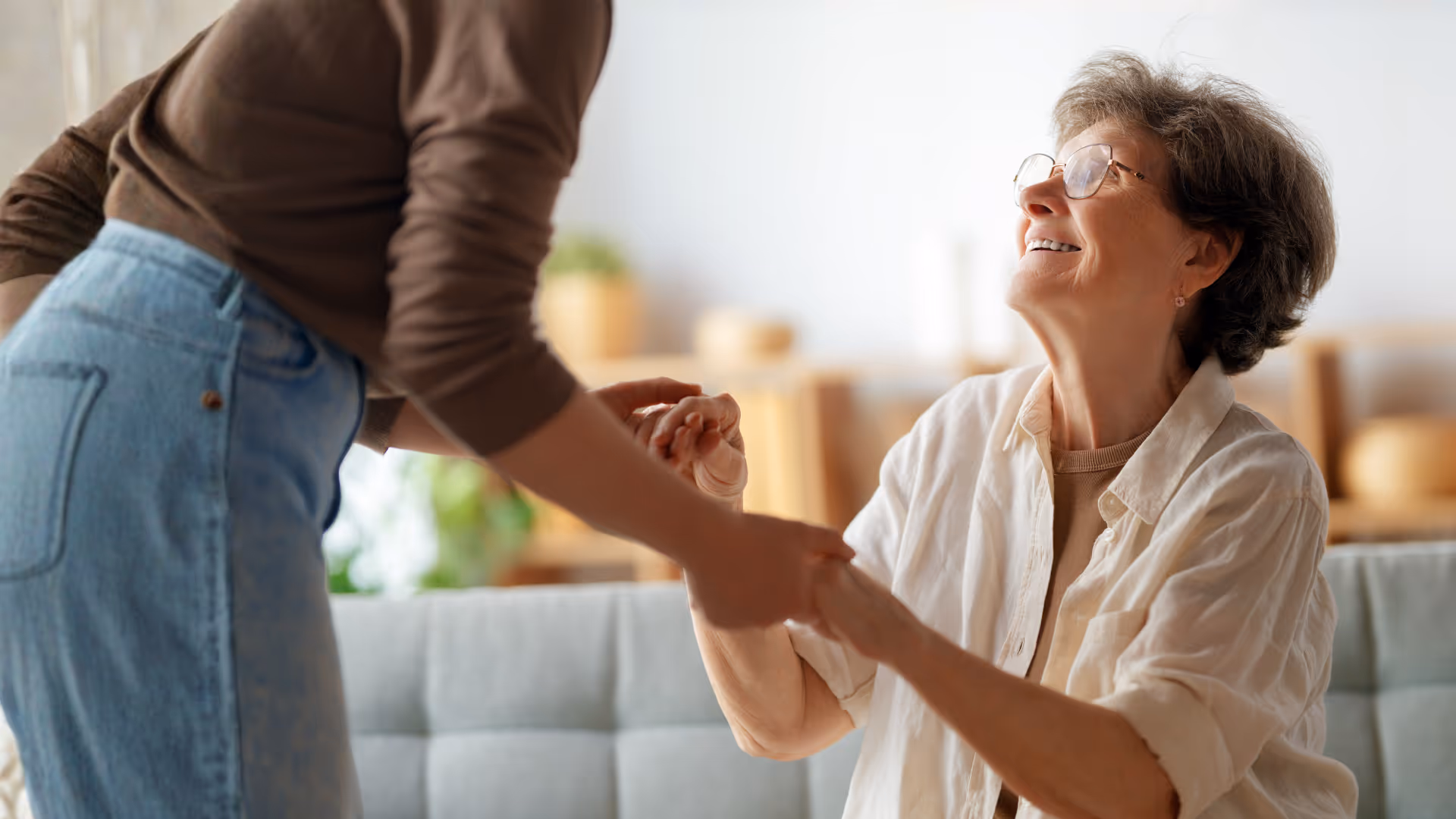Smiling elderly woman with glasses holding hands with a younger person in a cozy living room.