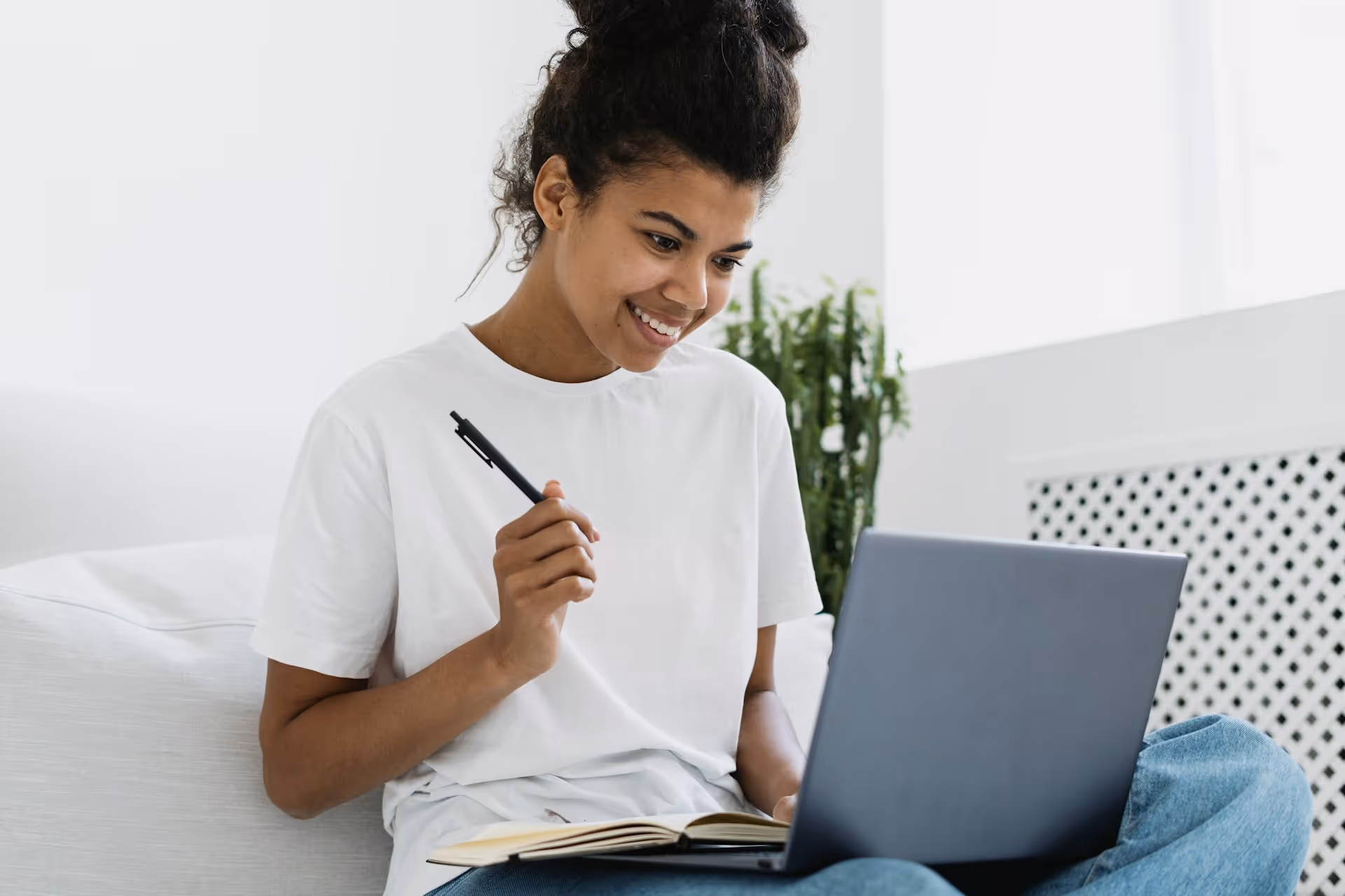 Jeune femme souriante utilisant un ordinateur portable assise sur un canapé avec un carnet et un stylo à la main.