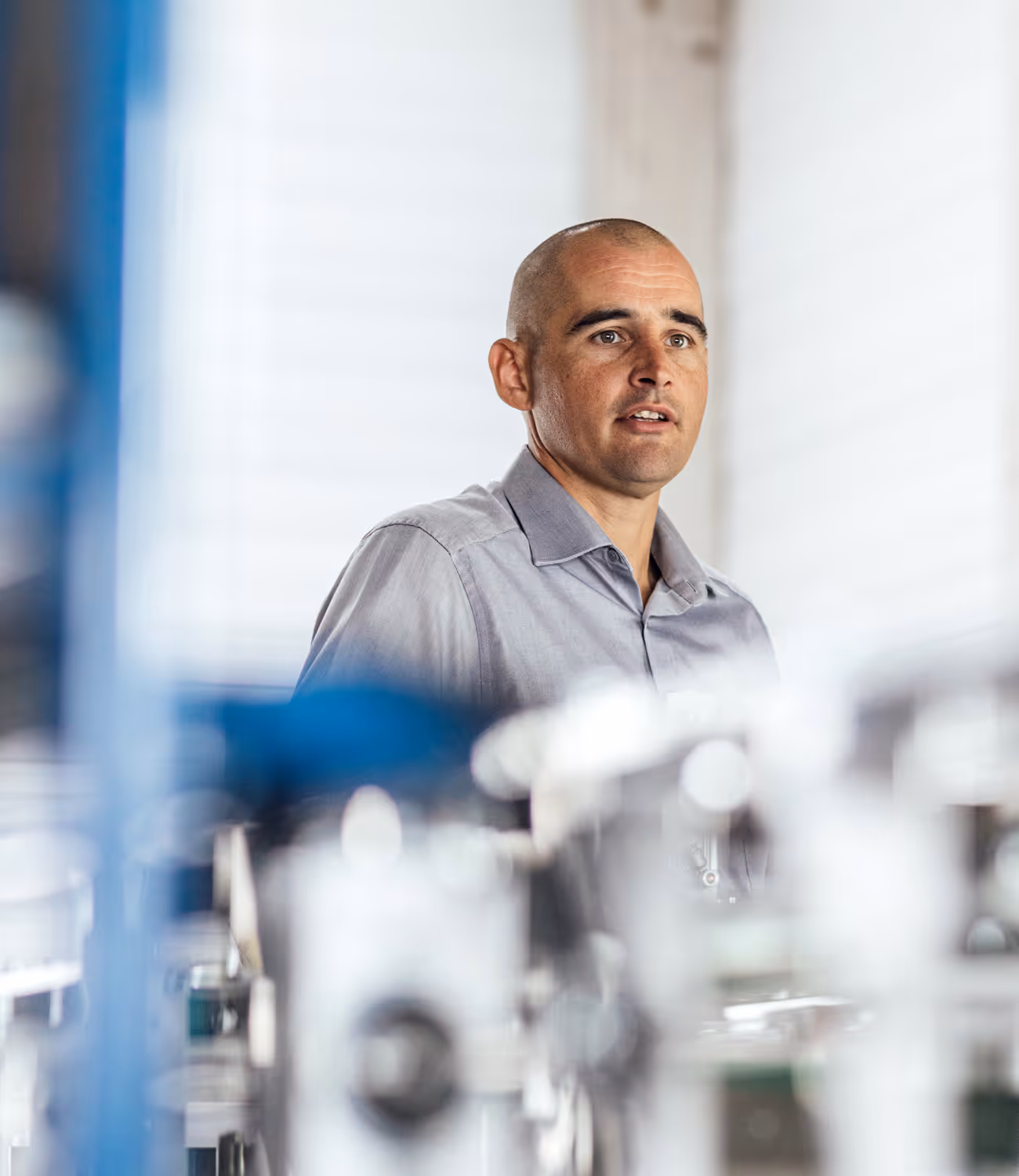 Portret van een man in een industriële omgeving, gekleed in een lichtgrijs hemd, met zachte scherptediepte en technische installaties op de voorgrond.