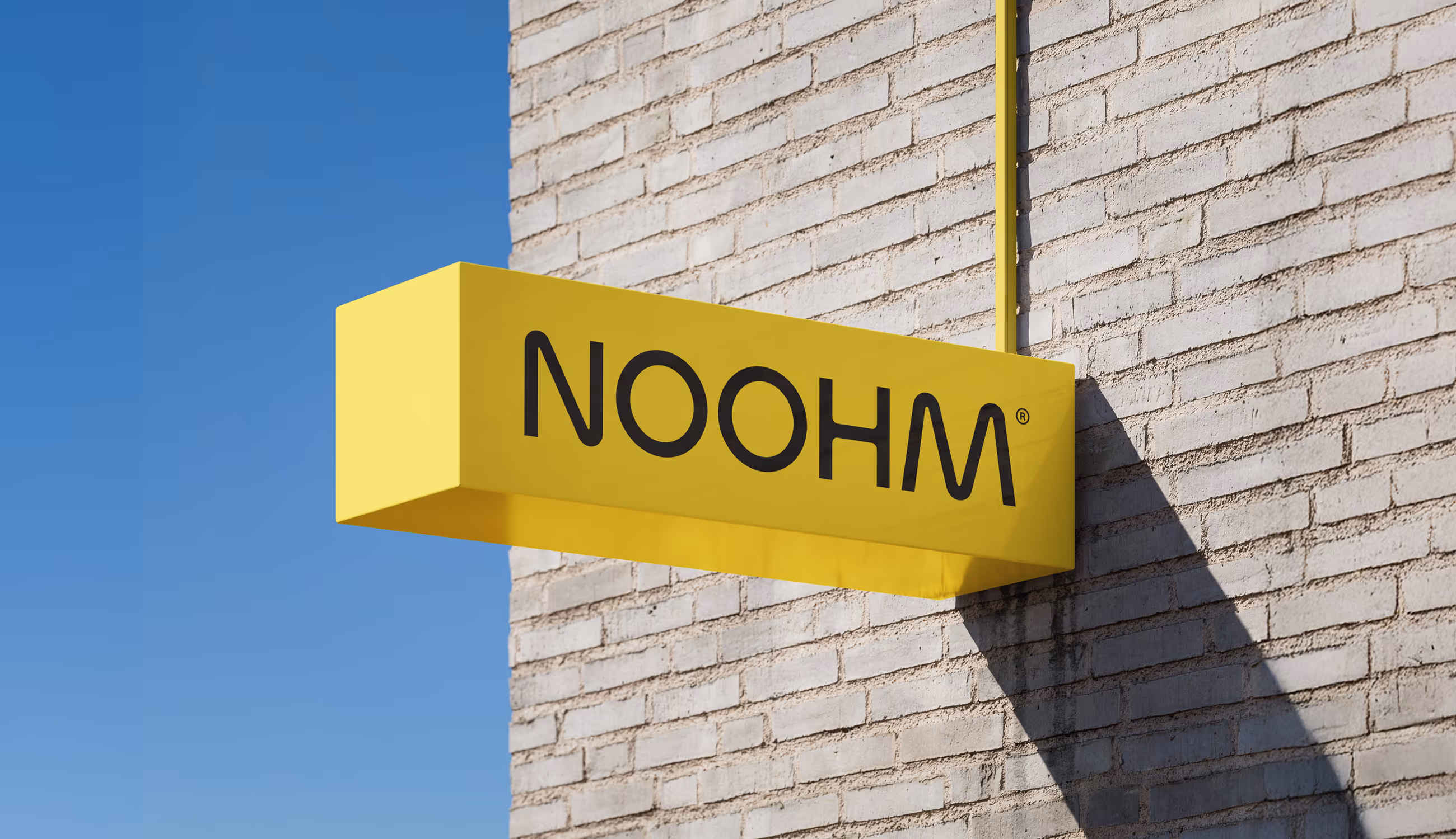 Gele verlichte gevelbox met zwart Noohm-logo, bevestigd aan een lichtgrijze bakstenen muur onder een heldere blauwe lucht.