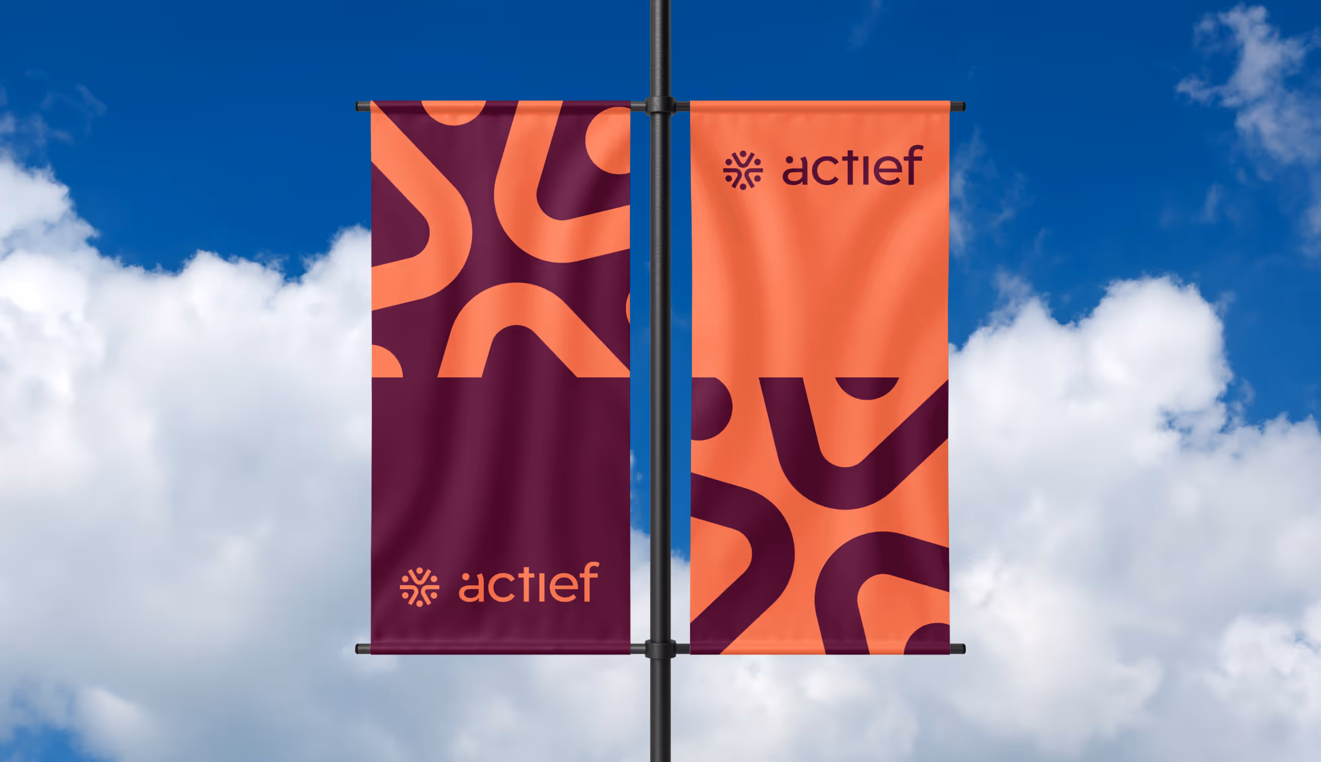 Twee verticale vlaggen aan een paal tegen een blauwe lucht, in oranje en paars met het Actief-logo en organische patroonvormen.