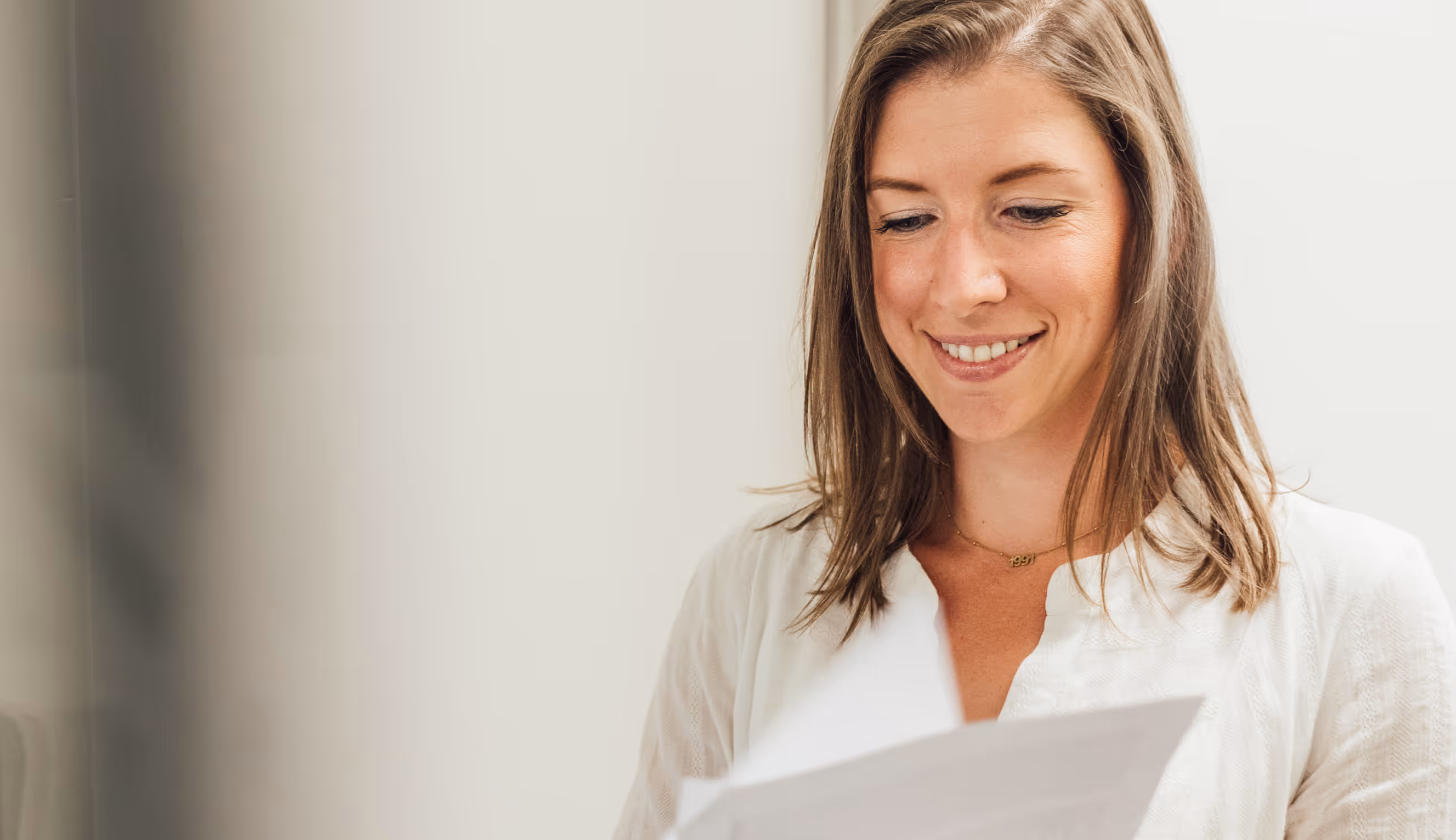 Vrouw in witte blouse die glimlachend naar documenten kijkt, in een lichte kantooromgeving.