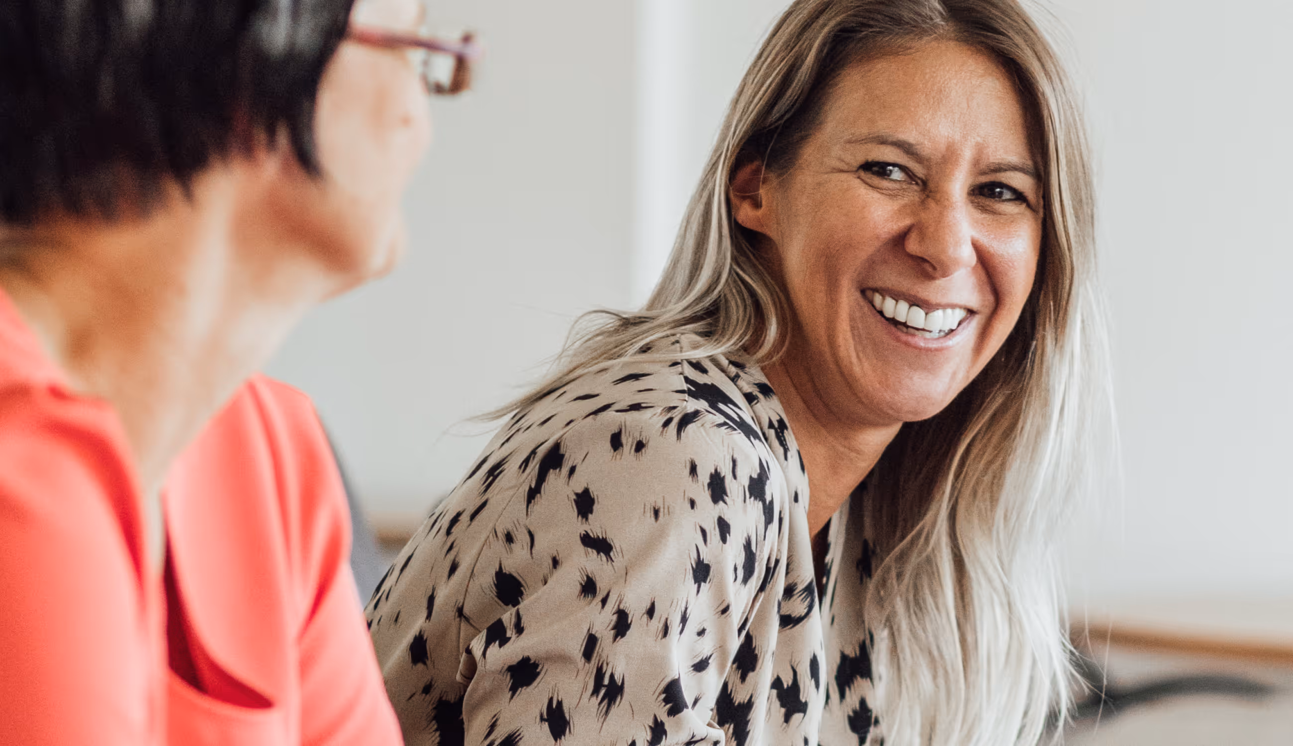 Twee vrouwen in gesprek aan een tafel, één met een beige blouse met zwart patroon, beiden lachend in een lichte kantoorruimte.