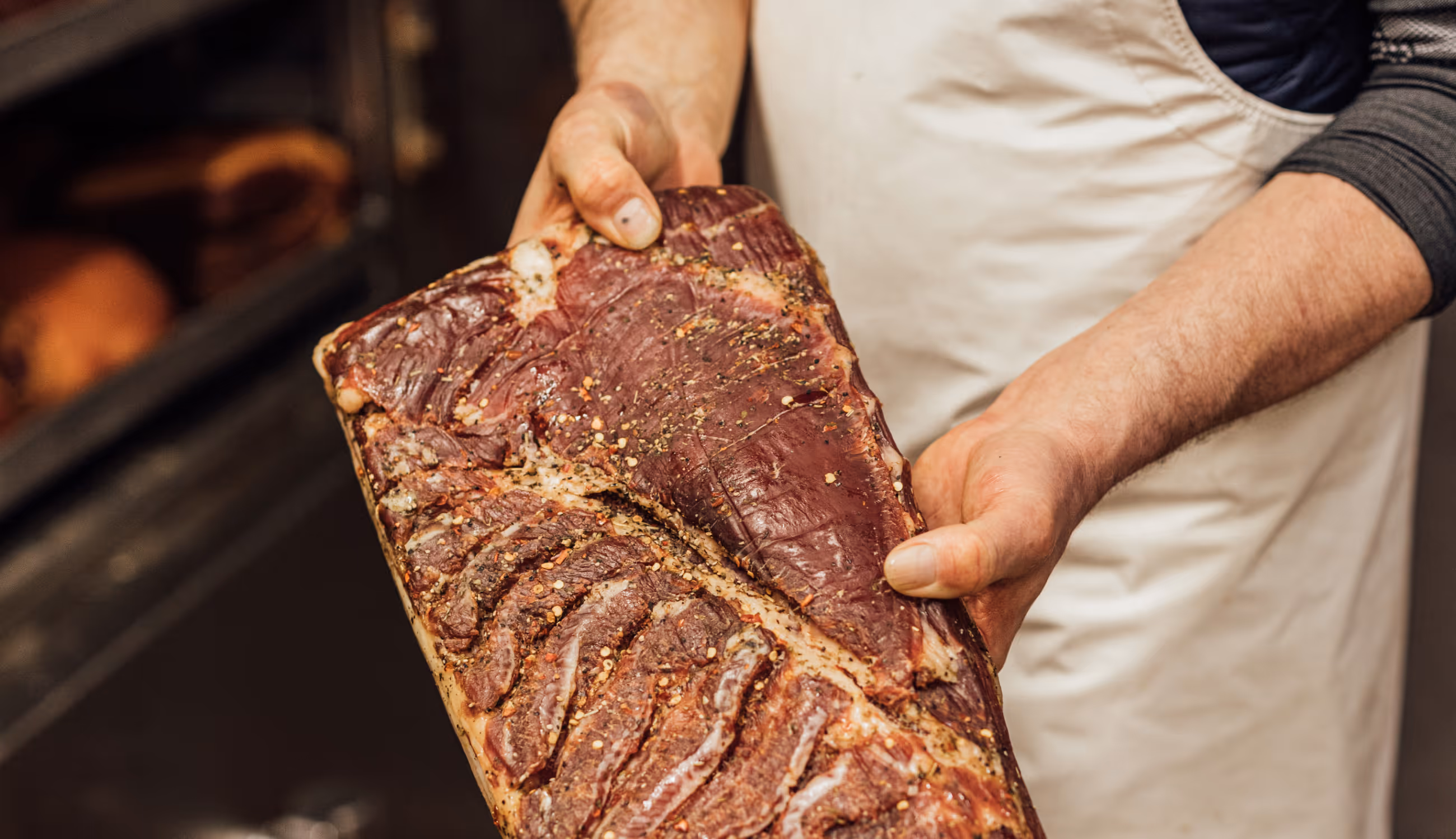 Close-up van handen die een gekruid stuk vlees vasthouden — ambacht, zorg en kwaliteit in één beeld.