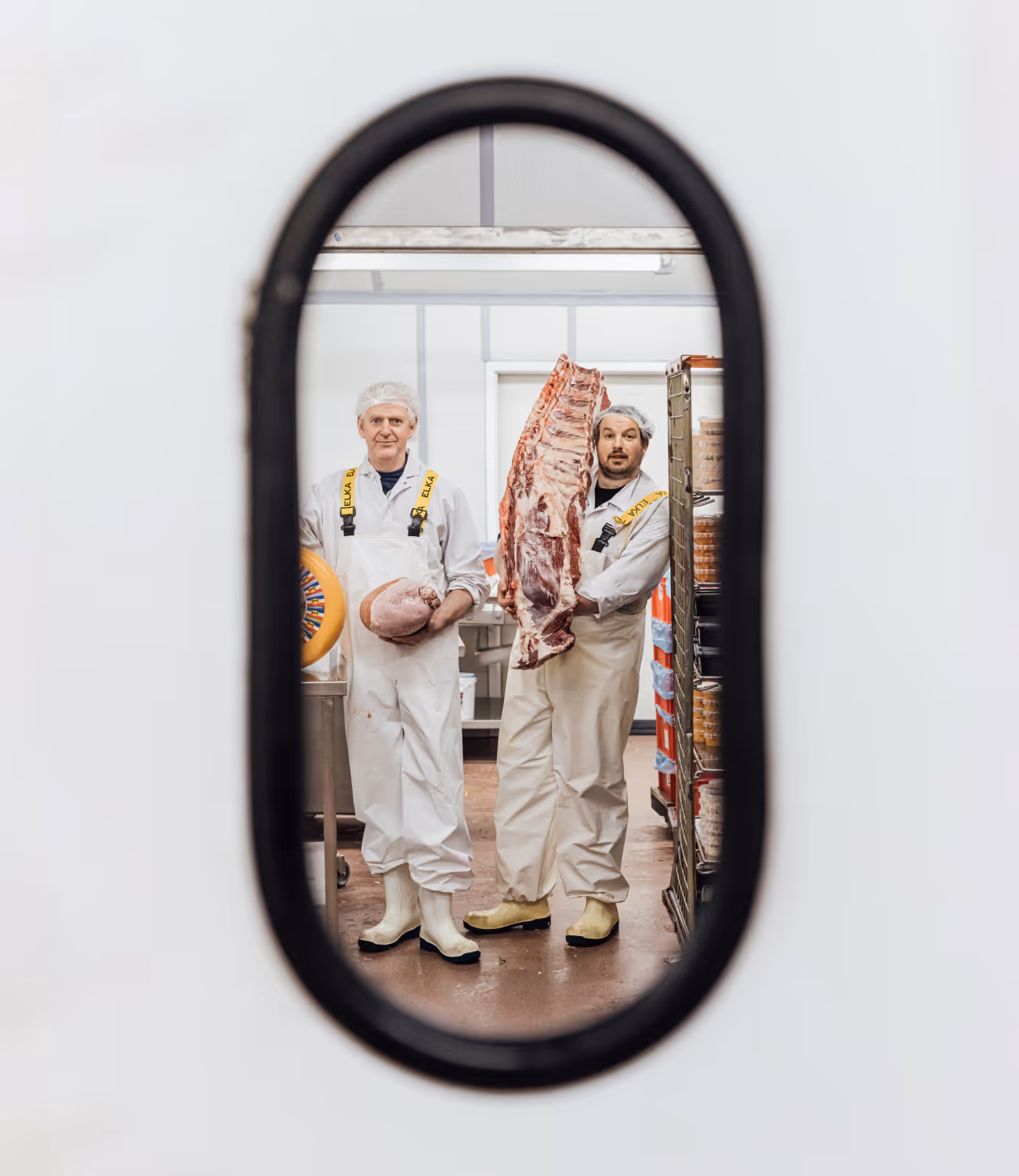 Twee medewerkers in witte werkkledij poseren met grote stukken vlees, gezien door een ovale deuropening.