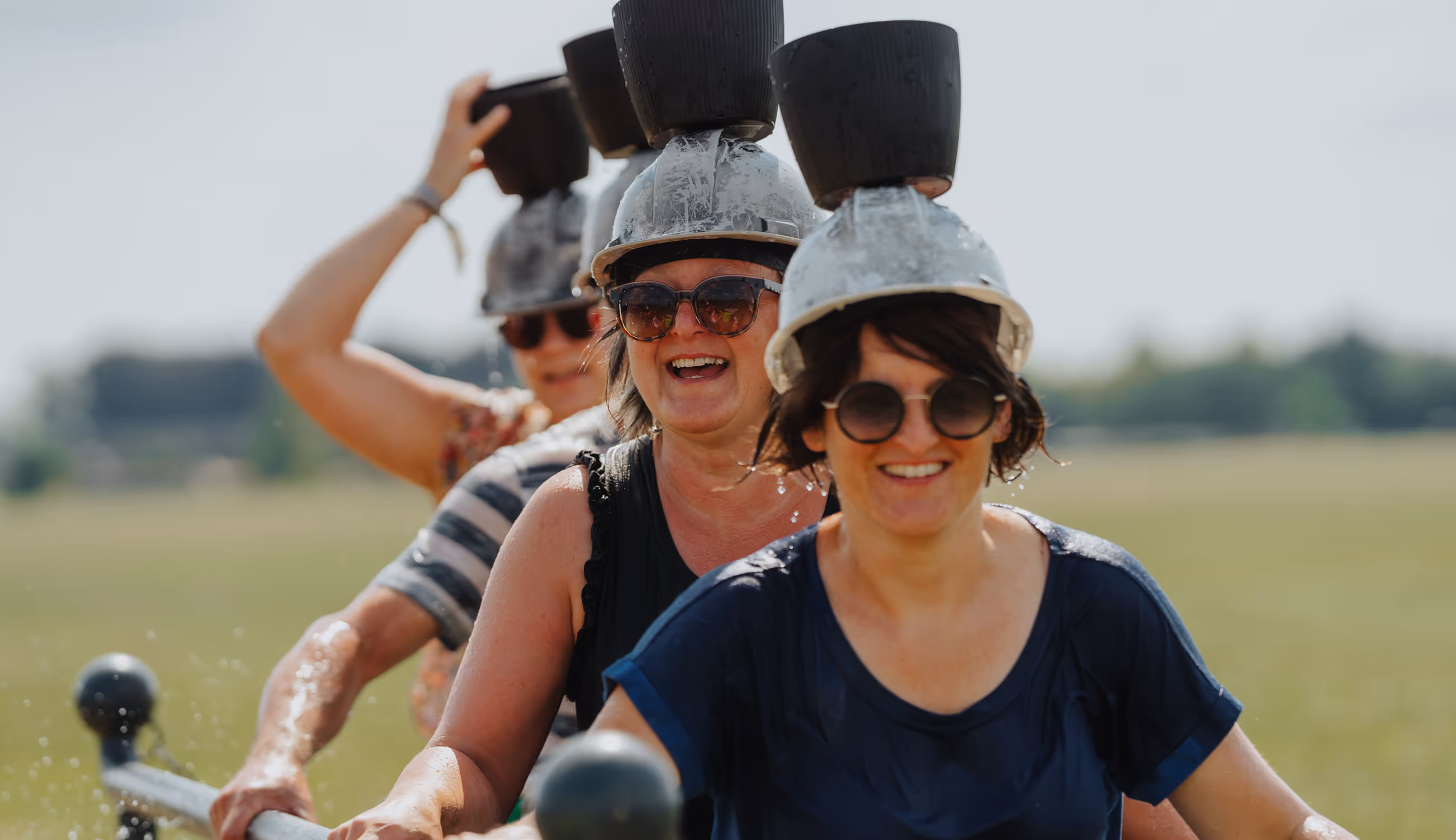 Vrouwen lachen tijdens een spel met emmers op het hoofd — zomers beeld vol fun en verbondenheid.