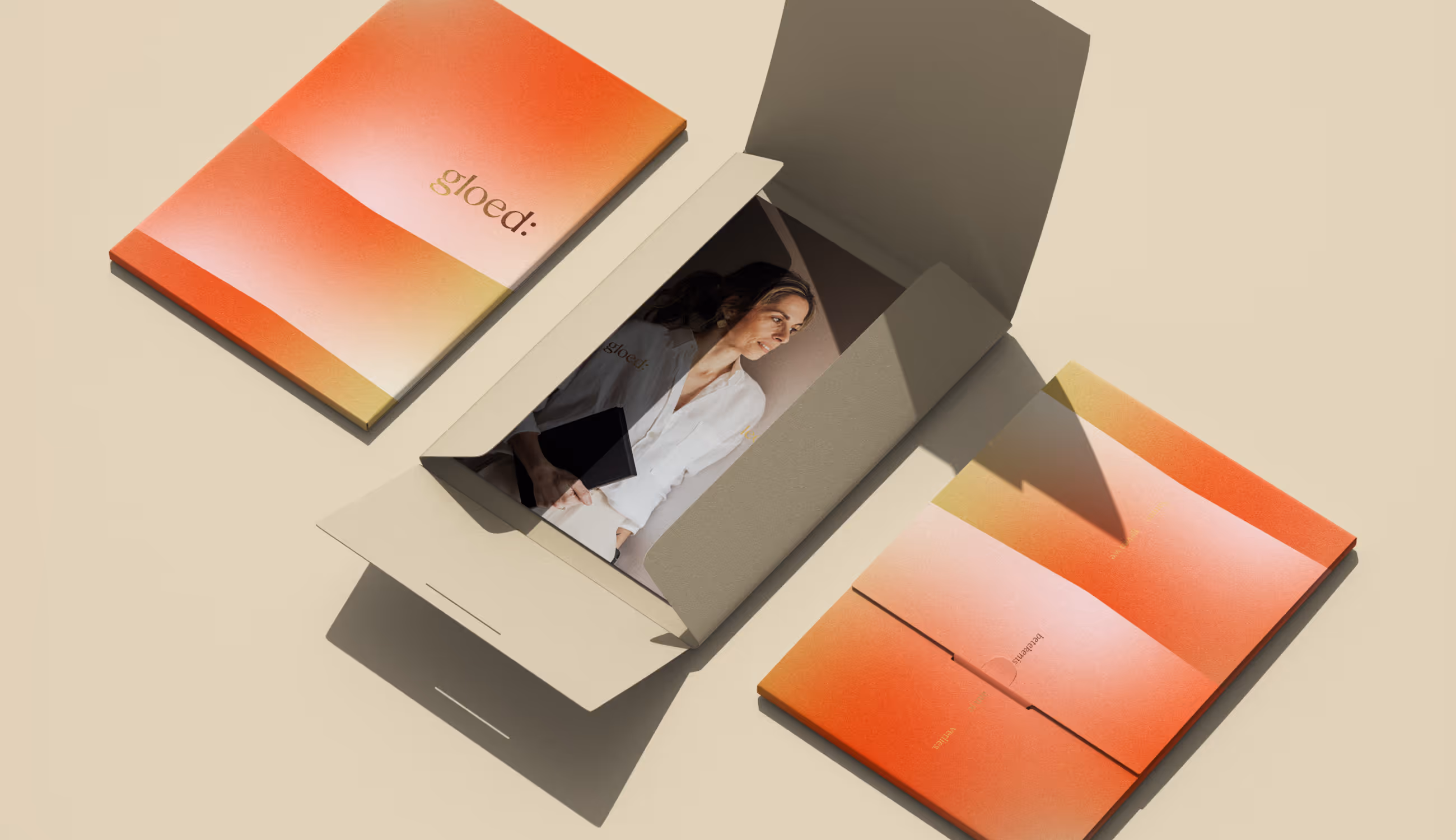 Een luxe, beige doos ligt open op een zachte zandkleurige ondergrond. Binnenin zie je een portret van een vrouw in witte blouse, terwijl rondom gradient mappen in warme oranje- en goudtinten liggen met het logo ‘gloed:’. Het beeld straalt warmte, zorg en verfijnde eenvoud uit.