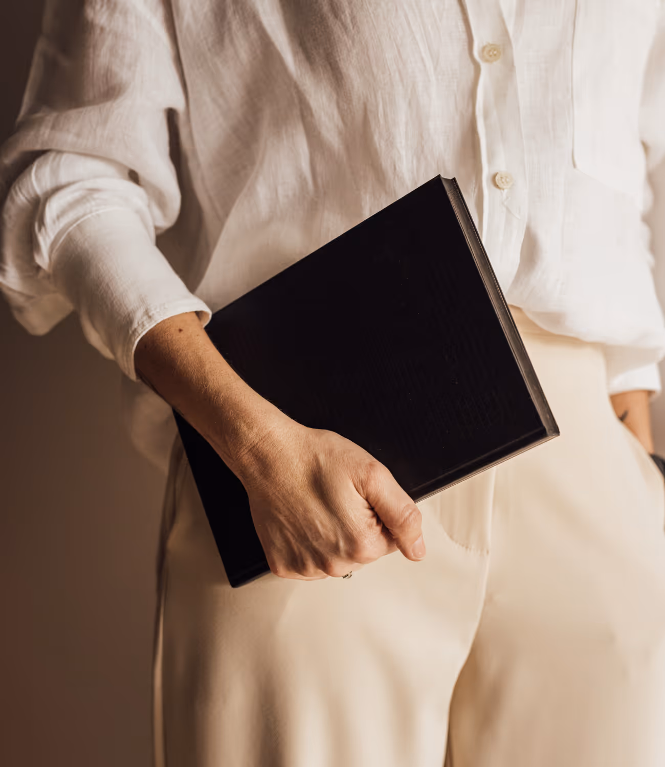 Close-up van een hand die een zwart boek vasthoudt tegen een lichte outfit. Het beeld toont rust, vertrouwen en concentratie.