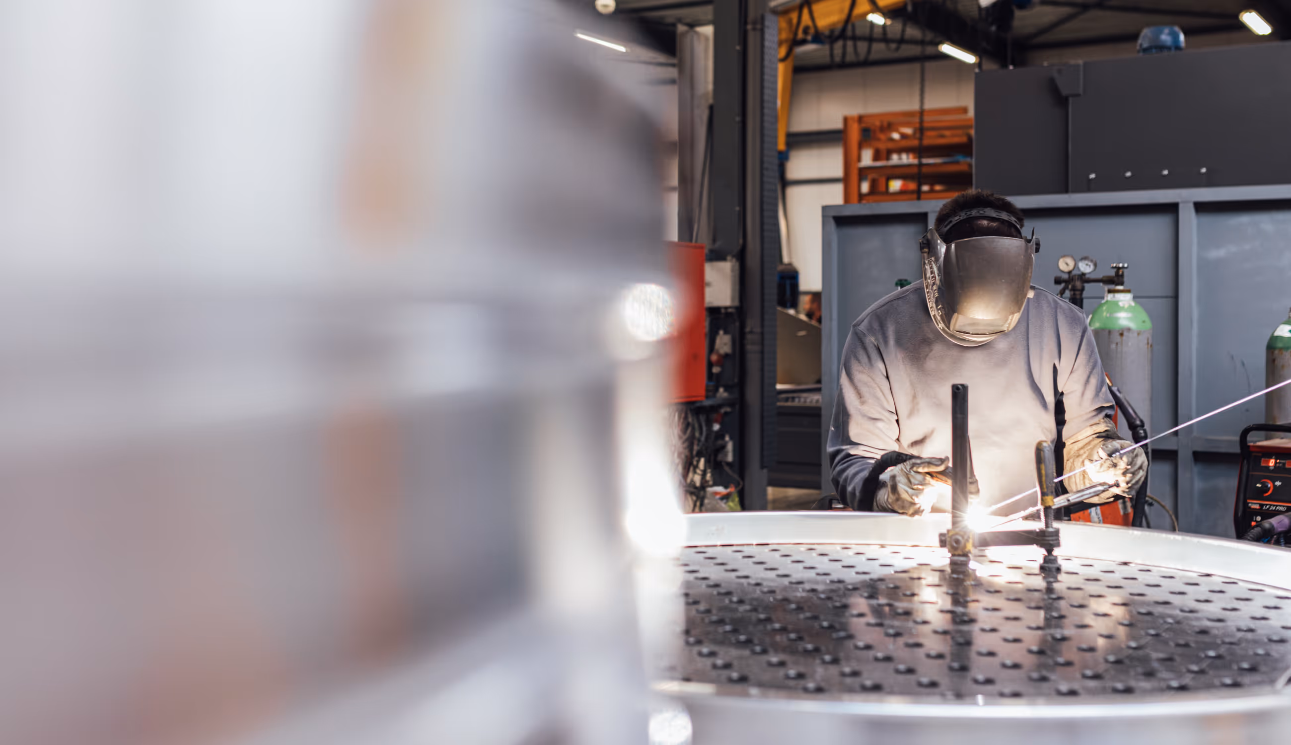 Een medewerker met lashelm last aan een ronde metalen constructie. De vonken en lichtreflecties creëren een industriële sfeer.