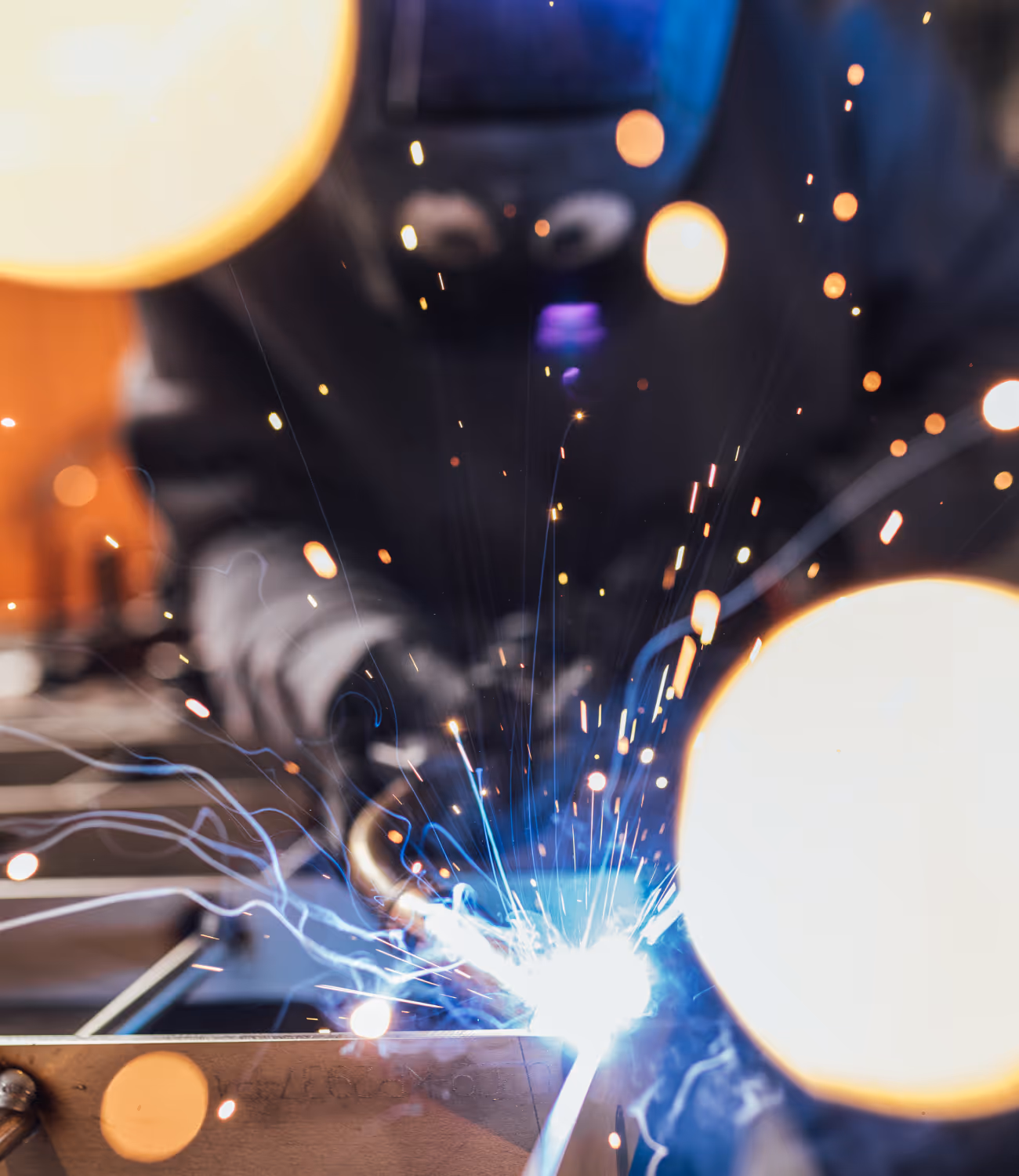 Close-up van een lassende medewerker met lashelm. Blauwe en oranje lichtvonken spatten in beeld, wat energie en vakmanschap oproept.