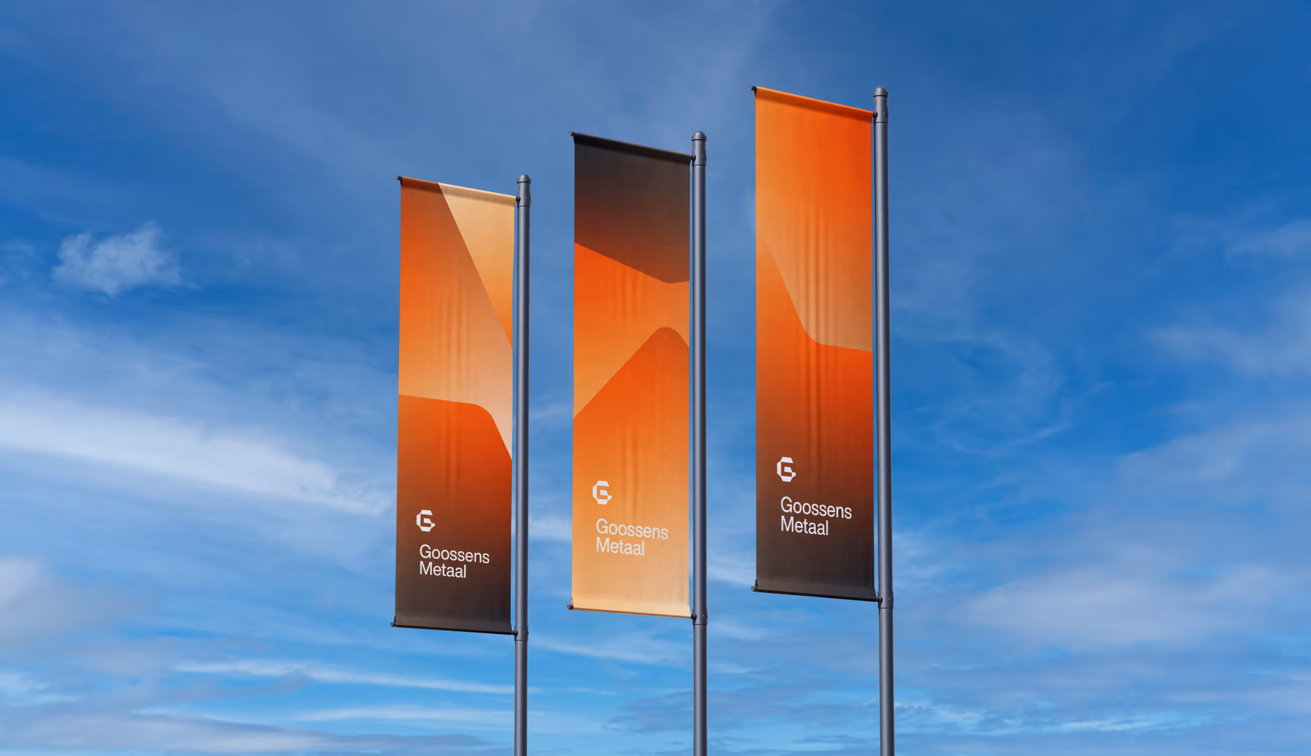 Drie oranje vlaggen met het logo van Goossens Metaal wapperen tegen een blauwe lucht. De grafische vlakken lopen in kleurverloop van oranje naar zwart.