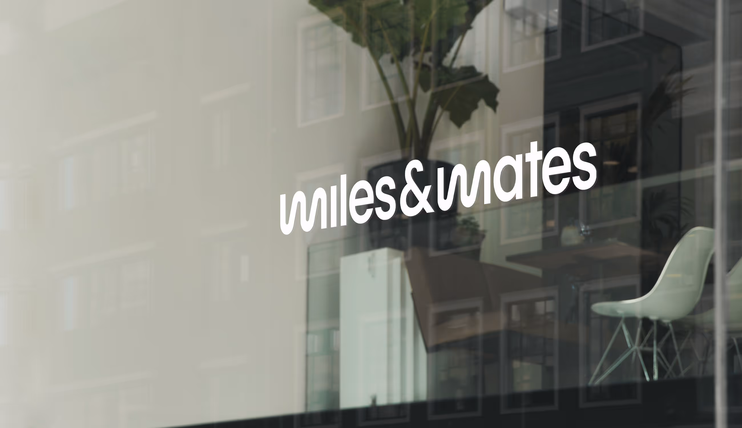 Wit Miles & Mates-logo op een glazen gevelraam, met zachte reflectie van de straat en een moderne kantoorruimte binnenin.