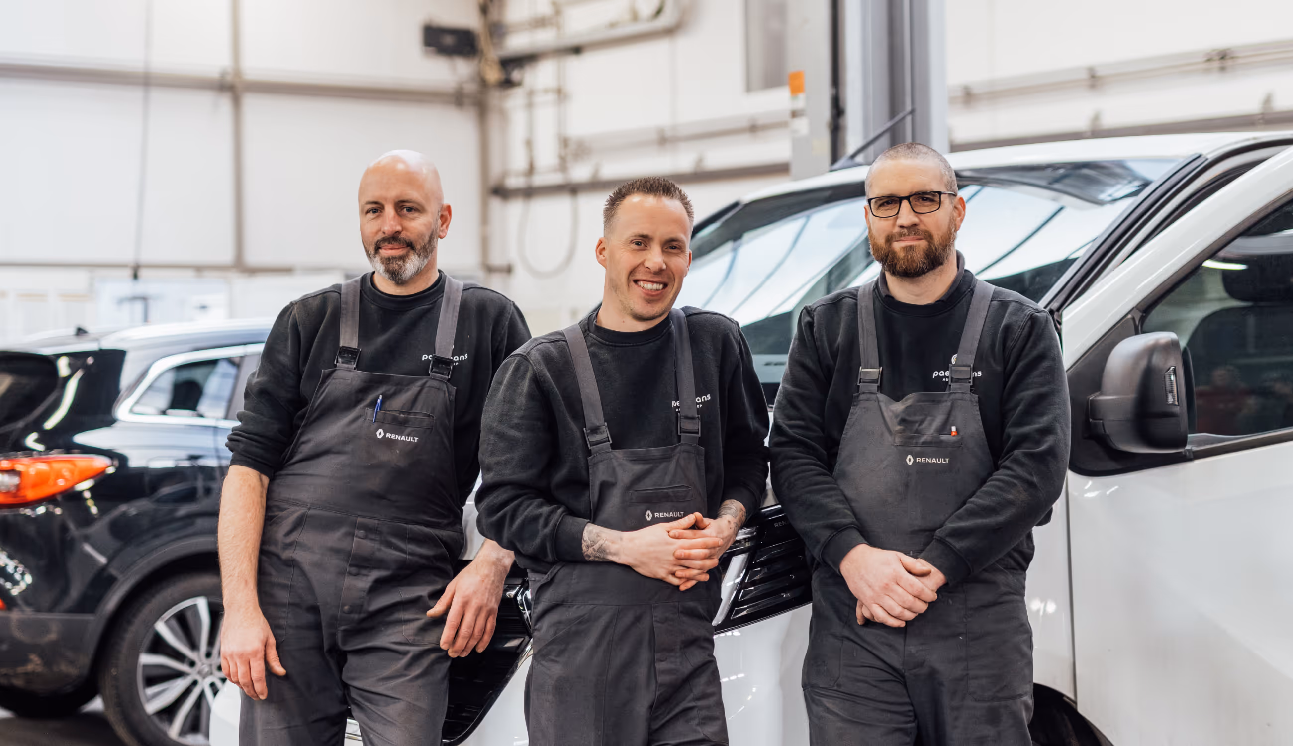 Drie monteurs in zwarte werkkledij poseren lachend in de garage tussen Renault-voertuigen.