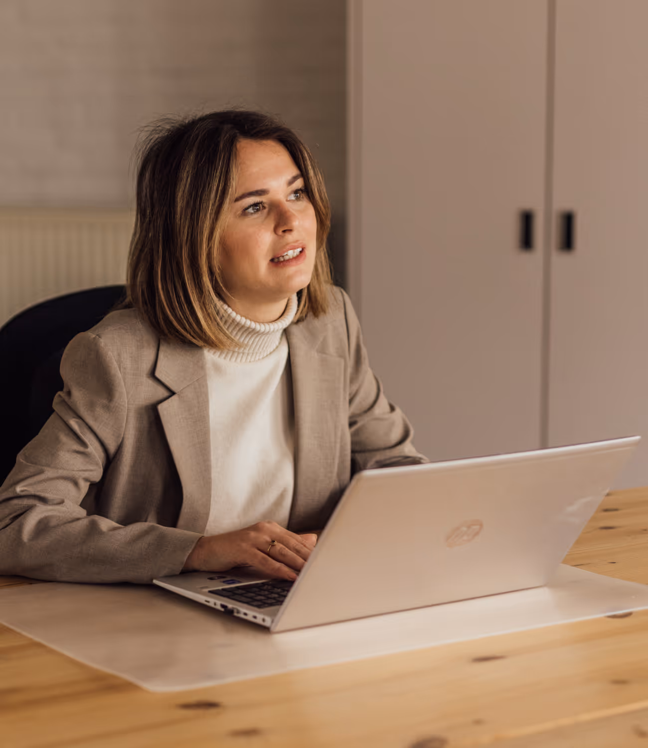 Vrouw in beige blazer en witte coltrui werkt geconcentreerd aan een laptop aan een houten tafel in een lichte kantoorruimte.