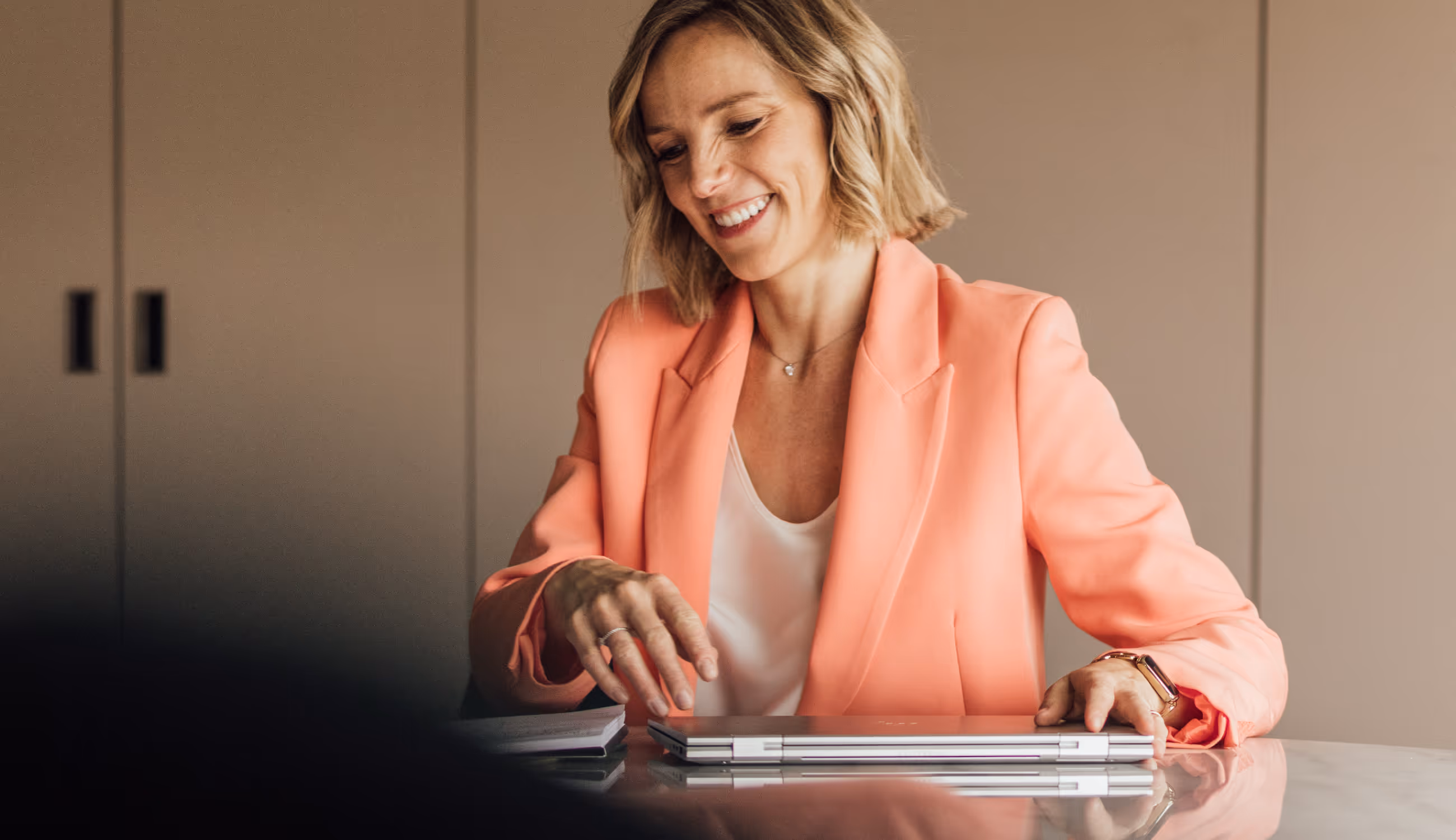 Vrouw in felroze blazer en witte top glimlacht terwijl ze aan tafel zit met een gesloten laptop.