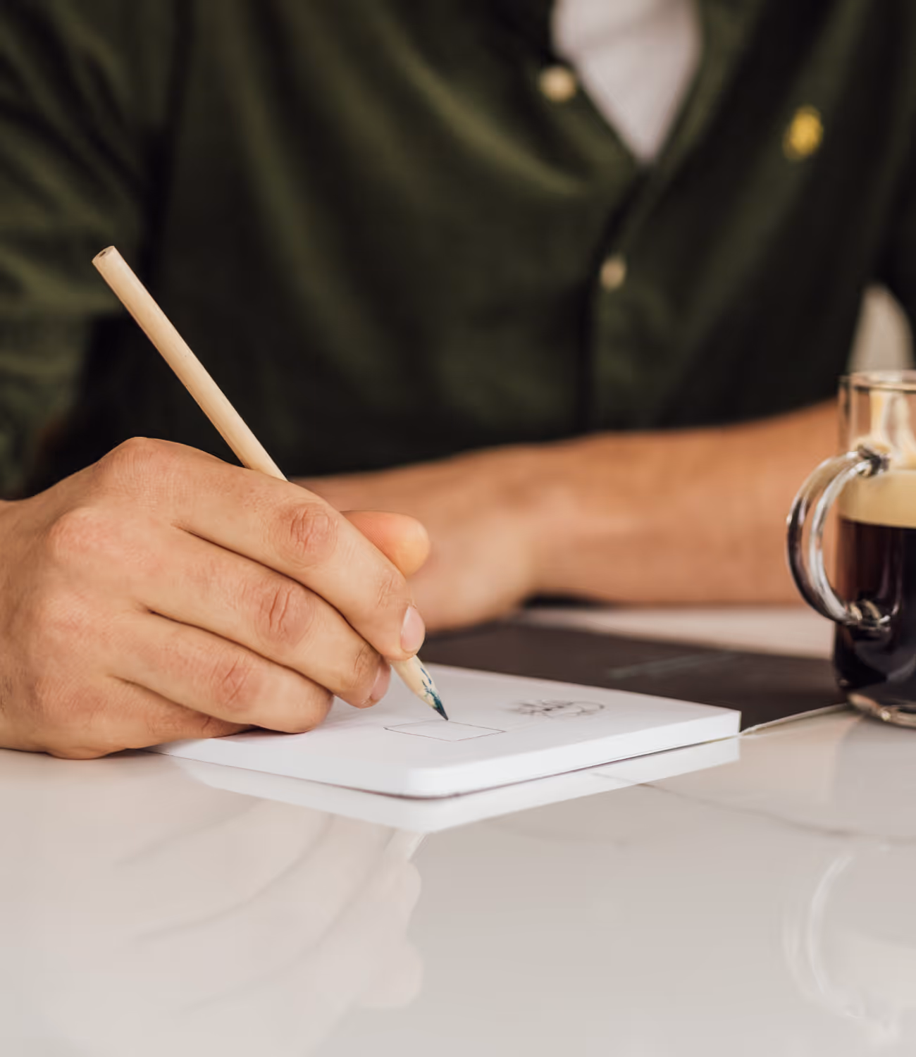 Close-up van een hand die schrijft met potlood in een notitieboek, naast een glas espresso.