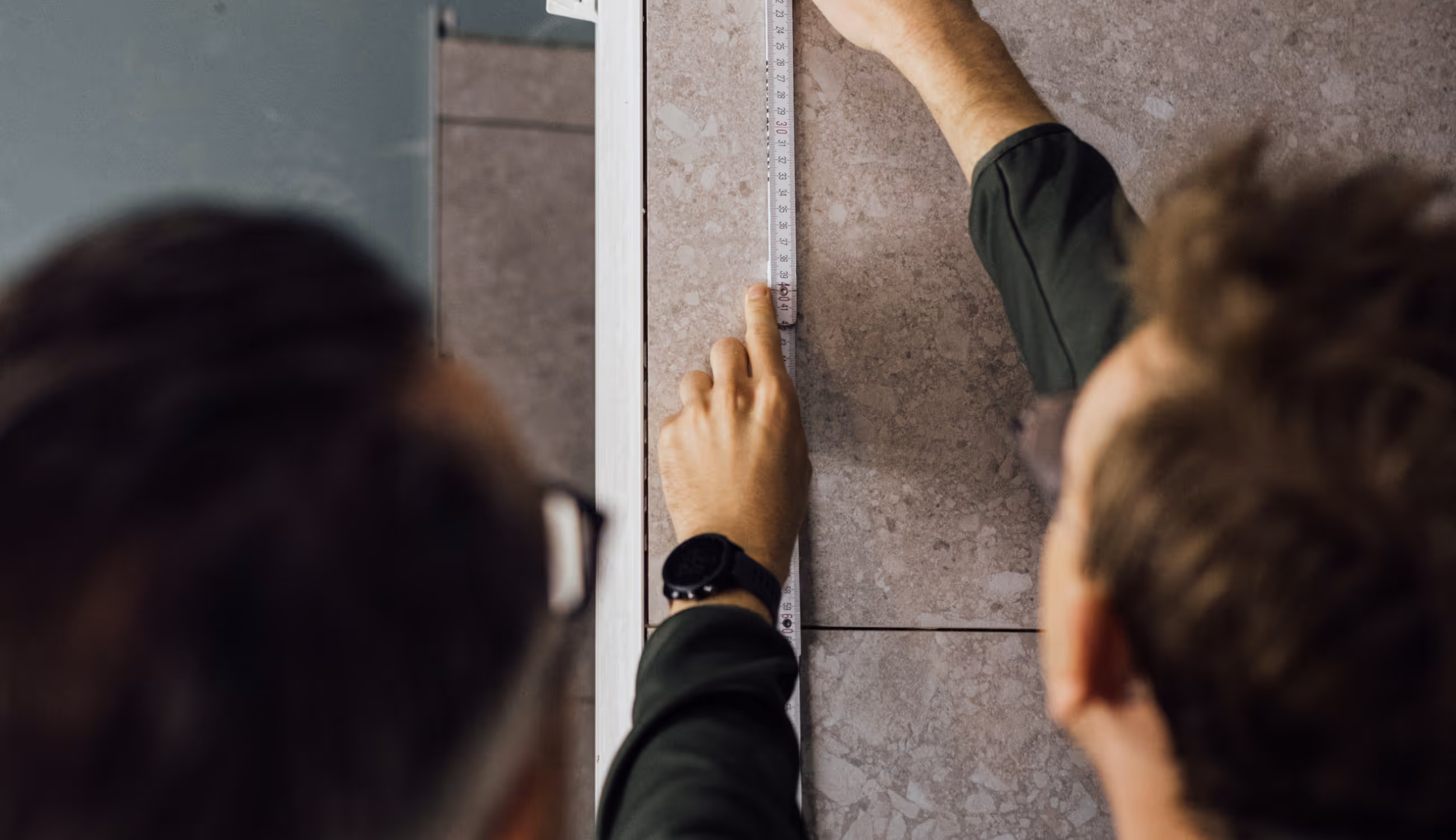 Twee mensen meten nauwkeurig met een meetlint tegen een betegelde wand, focus op handen en precisiewerk.