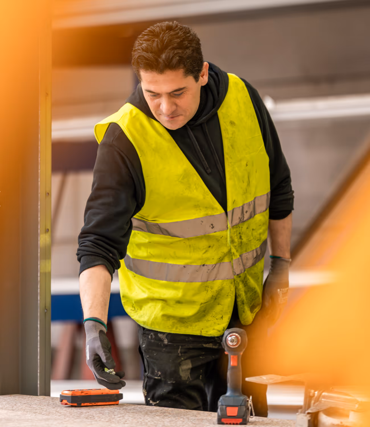 Werknemer in fluohesje plaatst gereedschap op werktafel, met warme lichtreflectie, symbool voor precisie en toewijding.