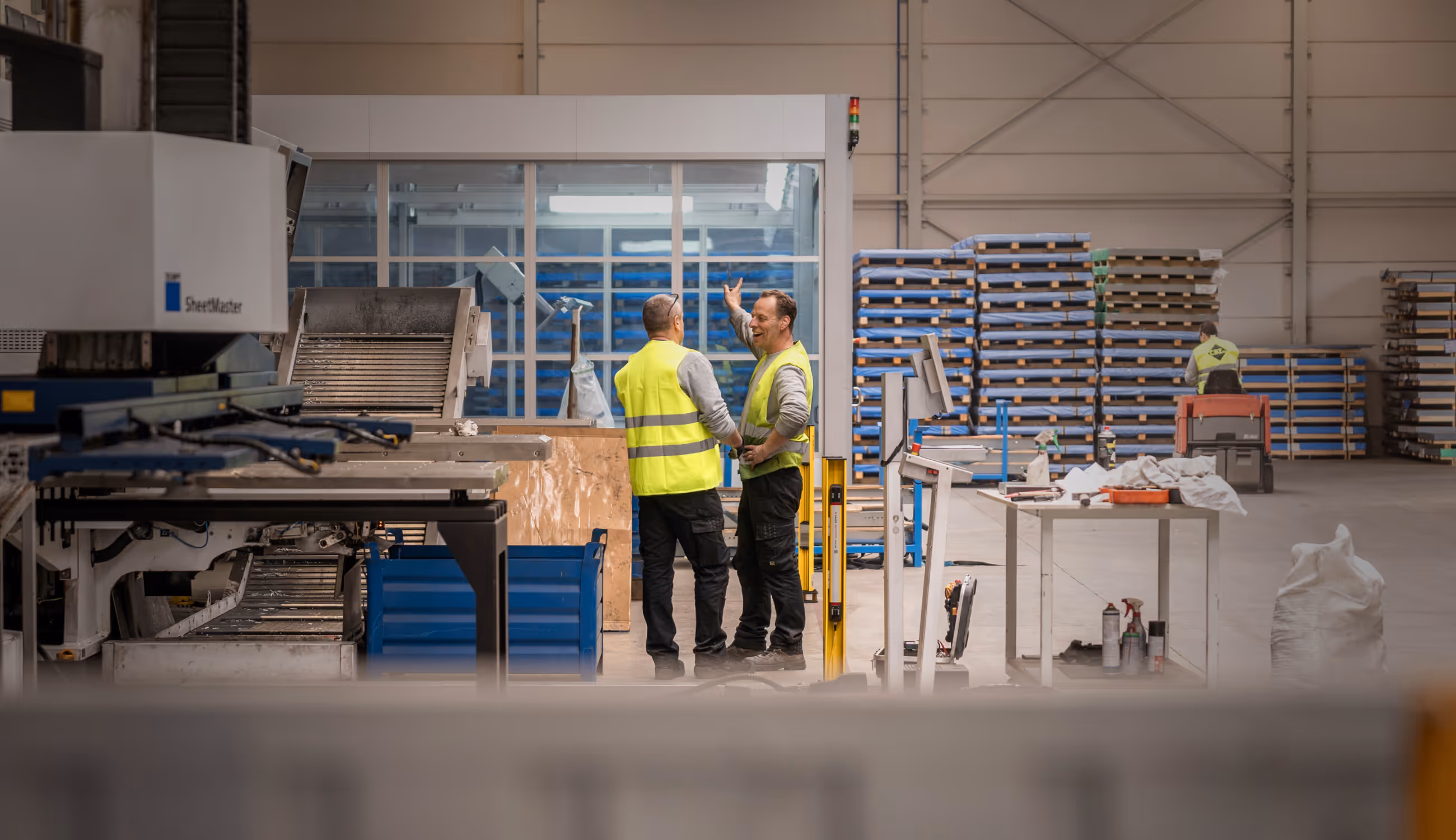 Twee Warsco-medewerkers praten lachend in een ruime fabriekshal, omringd door metalen platen en werktafels.