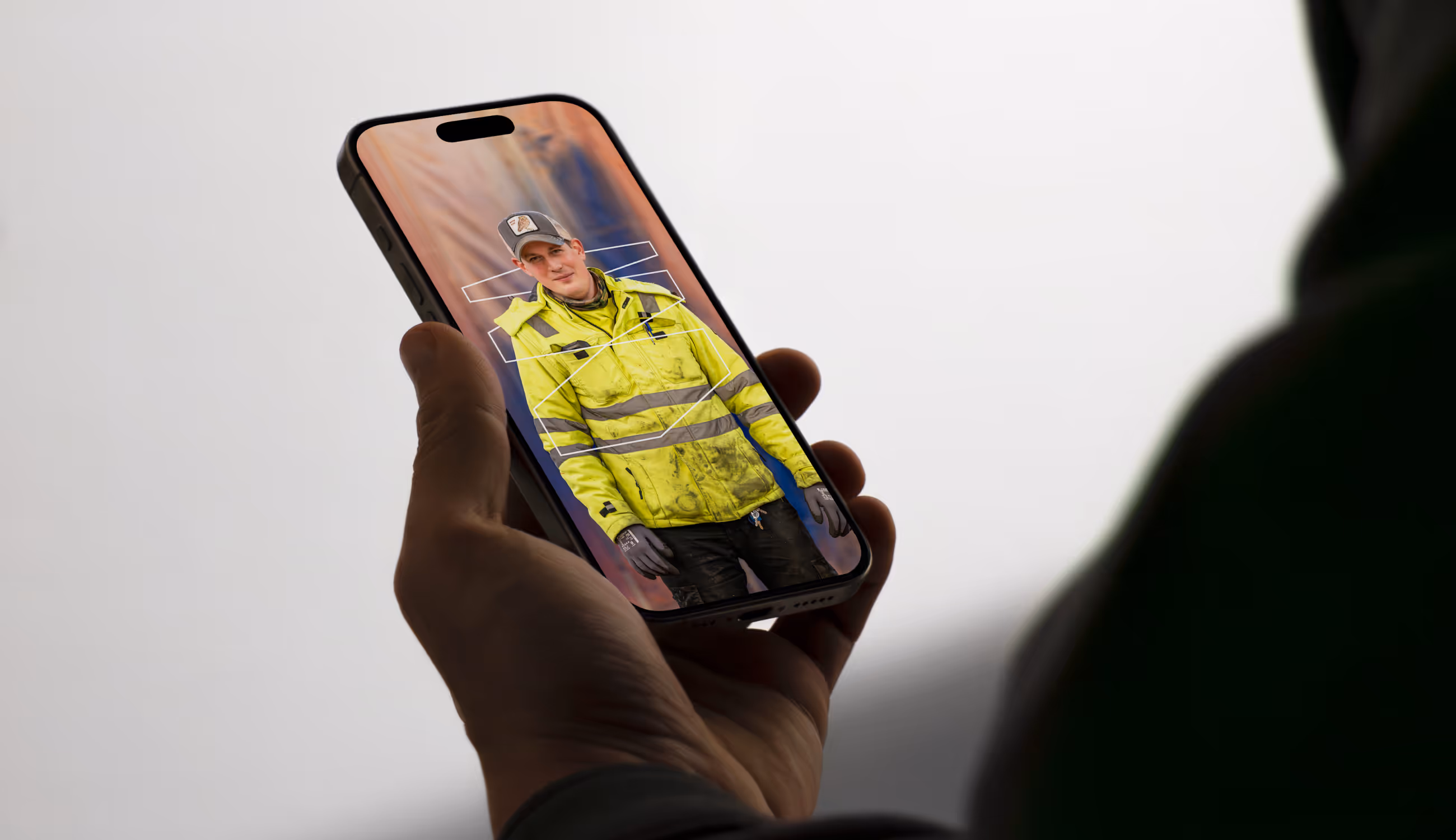 Hand houdt smartphone waarop een Warsco-medewerker in fluohesje en pet te zien is, tegen een zachte achtergrond van bouwkleuren, symbool voor de menselijke kant van vakmanschap.