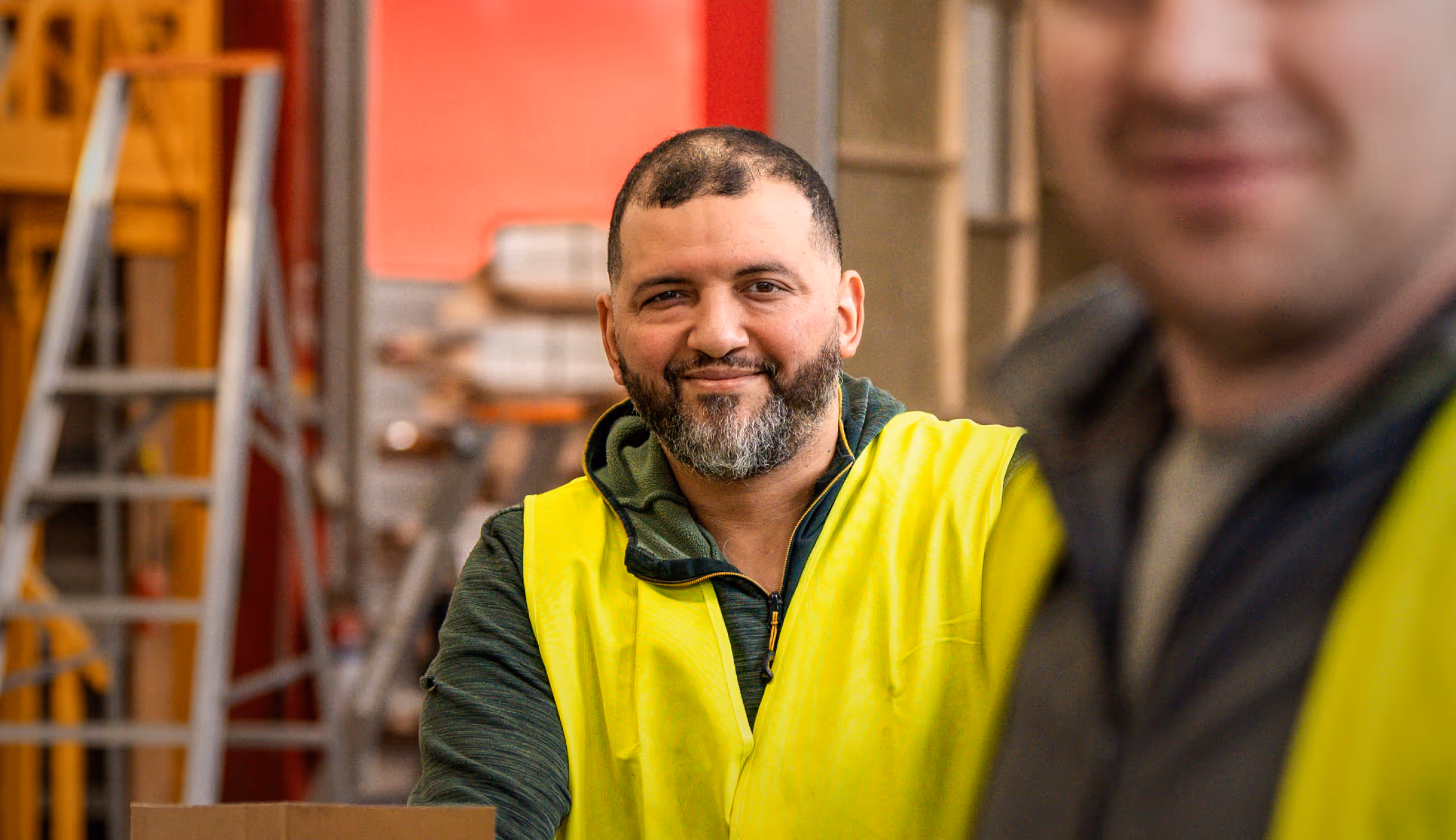 Warsco-medewerker met baard kijkt recht in de camera, op de achtergrond werkplaats en collega in vervaagde focus.