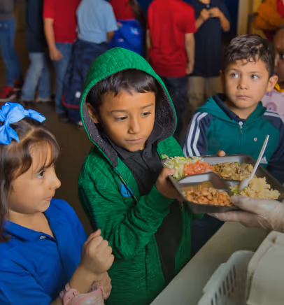 Three children receiving food on a tray from a person wearing gloves at a food distribution line.