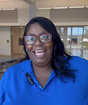 Smiling woman with glasses wearing a blue shirt standing indoors with tables and windows in the background.