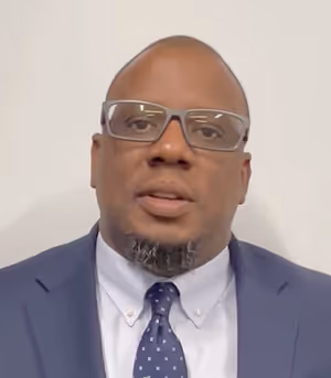 Man wearing glasses, a blue suit jacket, white shirt, and a blue polka-dot tie against a light background.
