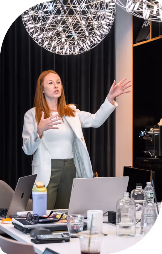 Woman in a white blazer speaking and gesturing with hands in a modern office meeting room with laptops and water bottles on the table.
