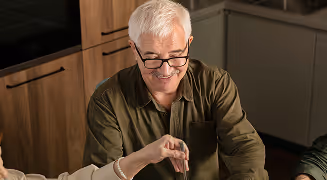 Smiling elderly man with white hair and glasses sitting inside and reaching out with his hand.