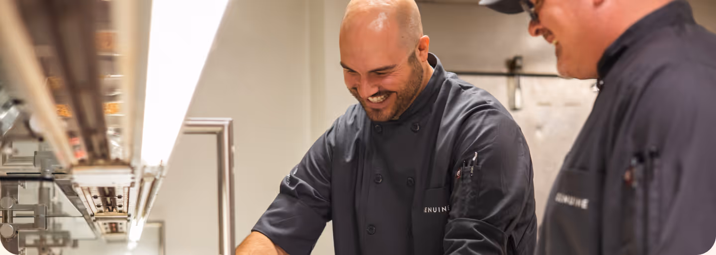 Two chefs in black uniforms smiling and working together in a kitchen.