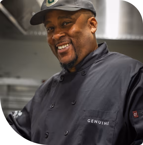 Smiling chef wearing a black uniform with the word 'GENUINE' on the chest pocket and a dark cap.