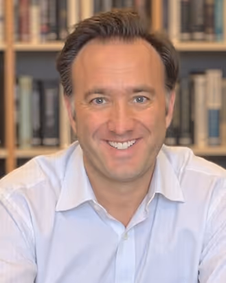 Smiling man with brown hair wearing a white collared shirt, sitting in front of a bookshelf.
