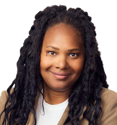 Smiling woman with long black dreadlocks wearing a beige jacket and white top, looking at the camera.