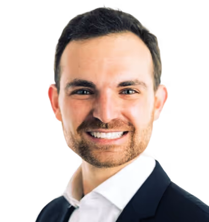 Smiling man with dark hair and beard wearing a white shirt and dark blazer against a transparent background.