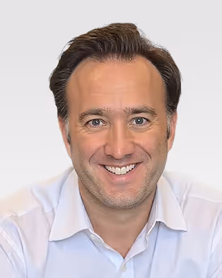 Smiling man with brown hair wearing a light blue collared shirt against a plain light background.