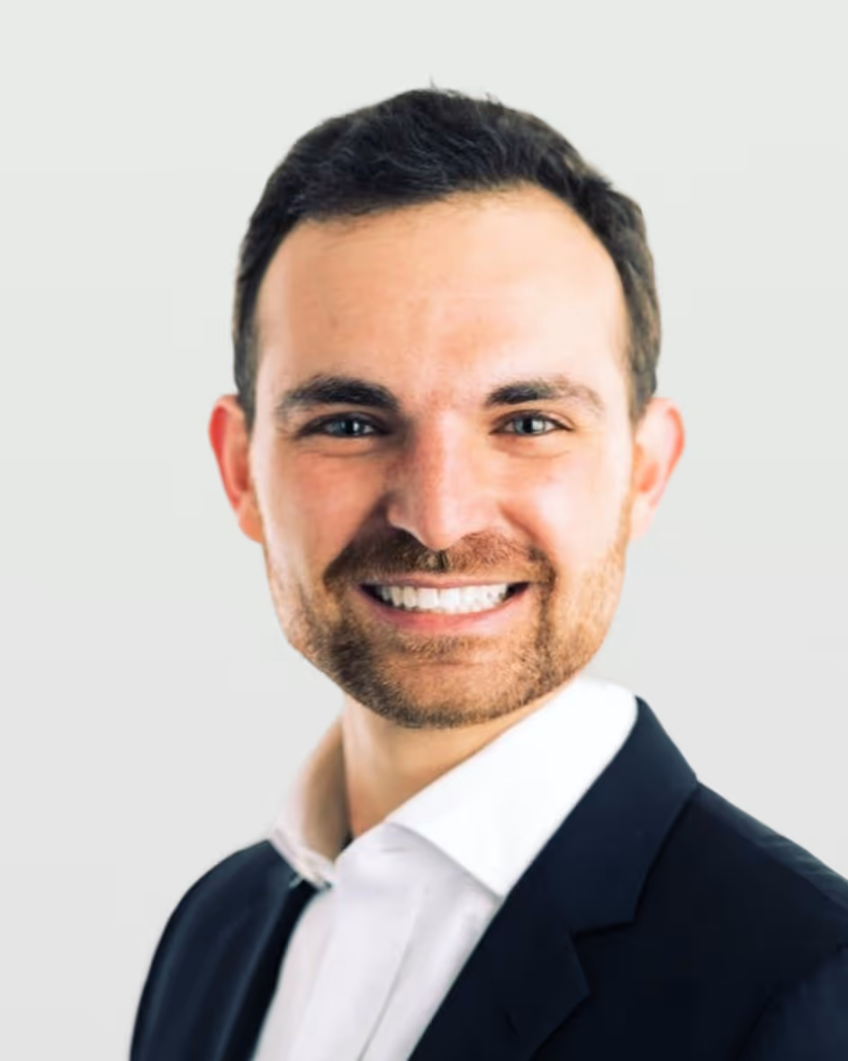 Smiling man with short dark hair and beard wearing a white shirt and dark blazer against a light background.