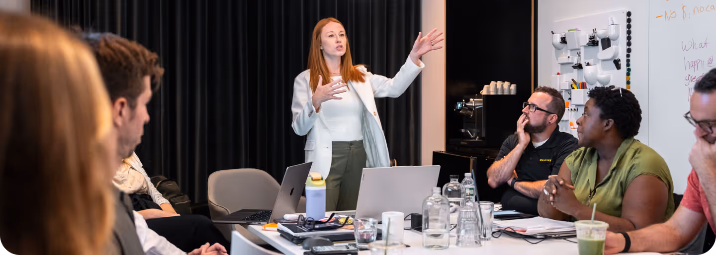 Woman in white blazer standing and speaking to a group seated around a conference table with laptops, notebooks, and drinks.