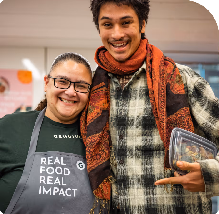 Smiling woman in glasses and apron with text 'Real Food Real Impact' standing next to smiling man wearing a scarf and plaid shirt, holding a food container.