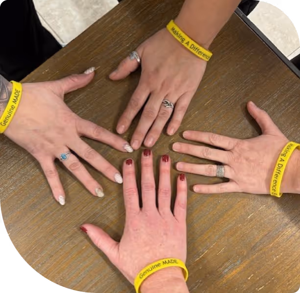 Four hands with rings and yellow wristbands reading 'Genuine MADE' and 'Making A Difference' placed on a wooden table.
