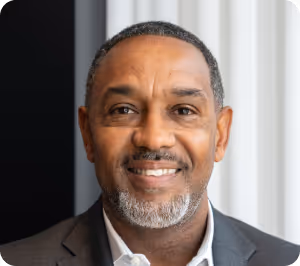 Smiling middle-aged man with short hair and gray beard wearing a dark blazer and white shirt in front of a window with curtains.