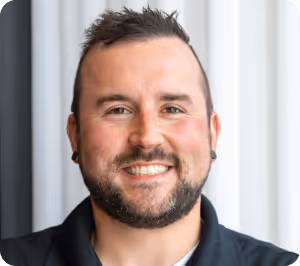 Smiling man with short dark hair and a beard wearing a black shirt in front of a light background.