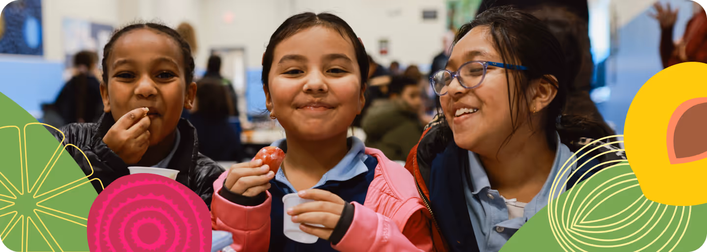 Three smiling girls sitting closely together, eating snacks and enjoying each other's company in a busy indoor setting.