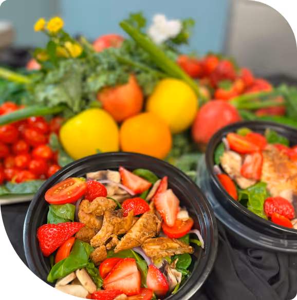 Two black bowls of fresh salad with grilled chicken, strawberries, cherry tomatoes, spinach, and mushrooms on a table with colorful fruits and vegetables in the background.