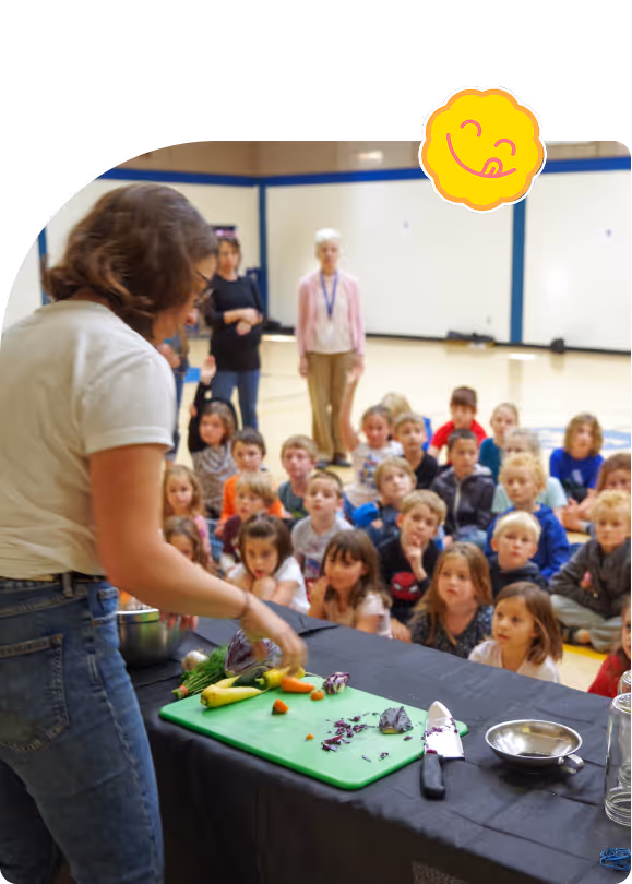 Person demonstrating vegetable preparation to a seated group of attentive children in a gymnasium.