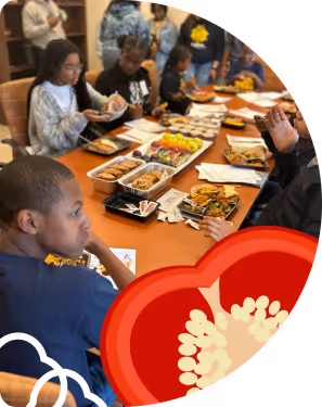 Several children seated around a table eating and sharing plates of food, with a decorative red heart graphic overlay on the corner.